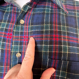 May include: Close-up of a plaid button-down shirt. The shirt features a classic plaid pattern in shades of blue, green, red, and yellow. The shirt has a collar, buttons, and a pocket. The fabric appears to be a soft flannel.