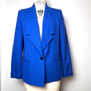 May include: A vibrant blue blazer with a single black button closure. The blazer features a notched lapel, two front pockets, and structured shoulders. The garment is displayed on a mannequin, showcasing its tailored fit and classic design.
