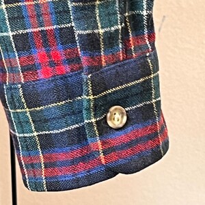 May include: Detail of a plaid fabric sleeve with a button. The tartan pattern includes red, blue, green, and yellow lines. The button is a light brown colour. The sleeve is part of a garment.