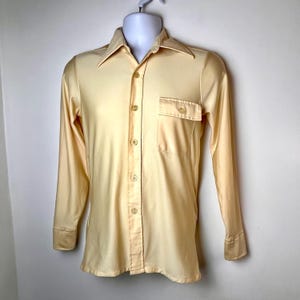 May include: A pale yellow long-sleeve shirt with a pointed collar, button-down front, and a single chest pocket. The shirt is displayed on a white mannequin and has buttoned cuffs.