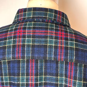 May include: A close-up shot of a plaid shirt's collar and upper back. The pattern is a grid of navy blue, green, red, and white lines. The shirt is made of a soft, textured fabric, and the collar is neatly constructed.