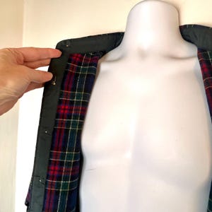 May include: A dark green jacket with a black collar and a red, blue, and green plaid lining. The jacket is displayed on a white mannequin. A hand is holding the jacket open.