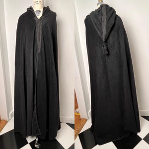 May include: A long, black hooded cloak made of a soft, velvety fabric. The cloak features a full-length design with a front zipper and a hood trimmed with a darker fabric. A tassel hangs from the back of the hood.