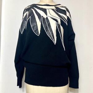 May include: Black sweater with a decorative shoulder design. The design features white and black leather-like shapes, outlined with white stitching and embellished with black beads. The sweater has long sleeves and a fitted waist.