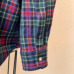 May include: Close-up of a blue, red, and green plaid flannel shirt sleeve. The cuff is buttoned with a silver button. The fabric has a soft, textured appearance.