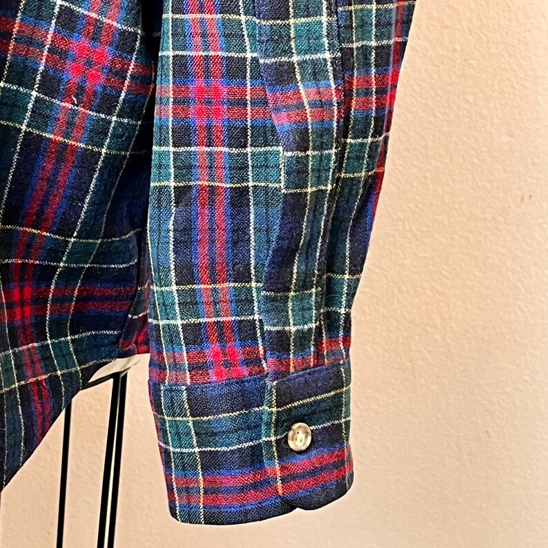 May include: Close-up of a blue, red, and green plaid flannel shirt sleeve. The cuff is buttoned with a silver button. The fabric has a soft, textured appearance.