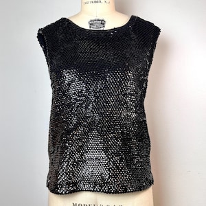 May include: A sleeveless black sequin top. The top is displayed on a mannequin. The sequins create a textured, shimmering effect. The neckline is rounded. The garment is a vintage piece.