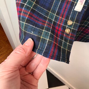 May include: Detail of a plaid jacket in navy blue, red, green, and yellow. Two tan buttons are visible on the fabric. The jacket's edge is held by a hand. The pattern is a classic tartan design.