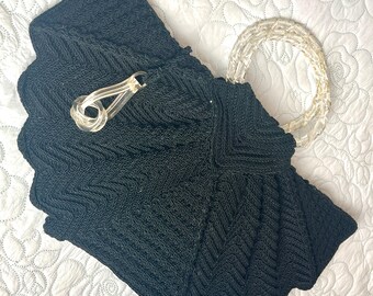 Puede incluir: Bolso de mano vintage negro con un cierre de plástico transparente y un asa trenzada. El bolso tiene un borde festoneado y un patrón texturizado en forma de chevrón. El asa está hecha de un material blanco tejido.
