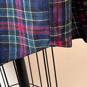 May include: Detail of a plaid fabric, showing a pattern of white lines on a dark blue base, with red and teal intersecting lines. The fabric is likely woven, possibly wool or a similar textile. The pattern is a classic tartan design.