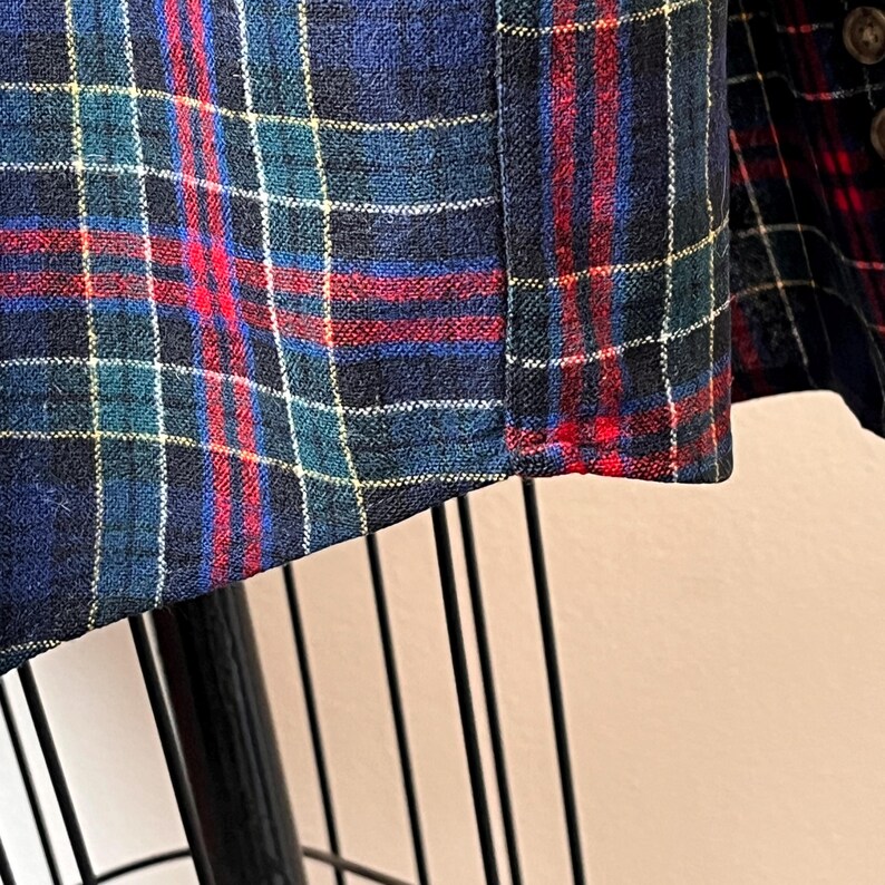 May include: Detail of a plaid fabric, showing a pattern of white lines on a dark blue base, with red and teal intersecting lines. The fabric is likely woven, possibly wool or a similar textile. The pattern is a classic tartan design.