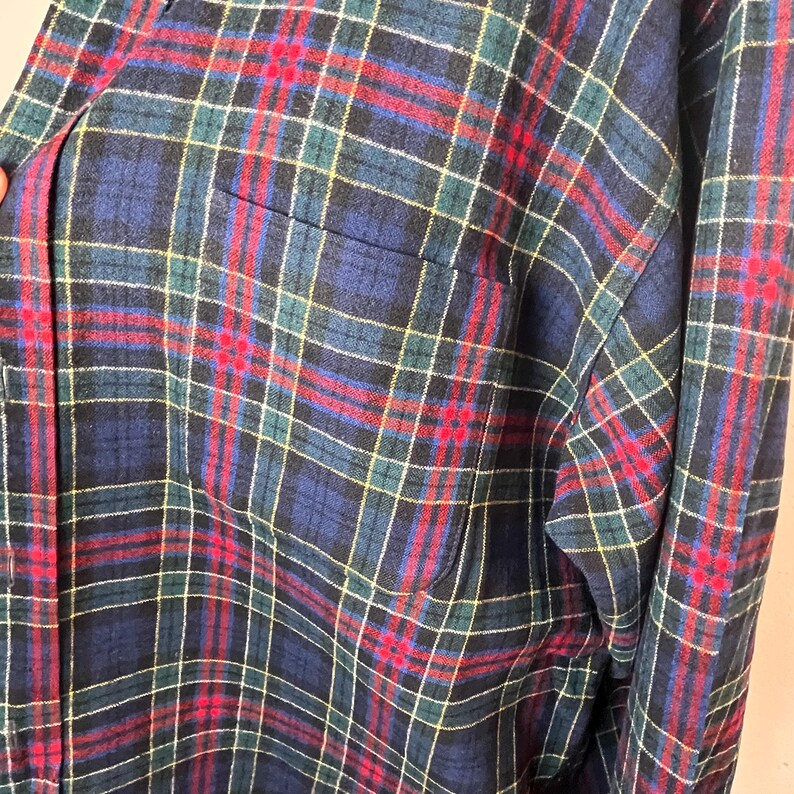 May include: Close-up of a plaid shirt in a traditional pattern. The shirt has a navy blue base with red, green, and yellow intersecting lines. A pocket is visible on the left. The fabric looks soft and comfortable.