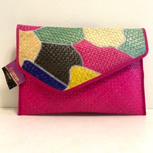 May include: A bright pink woven clutch with a patchwork flap. The flap features a geometric pattern of woven squares in various colors, including green, yellow, black, and blue. The tag reads "Carefree fashion spirit."