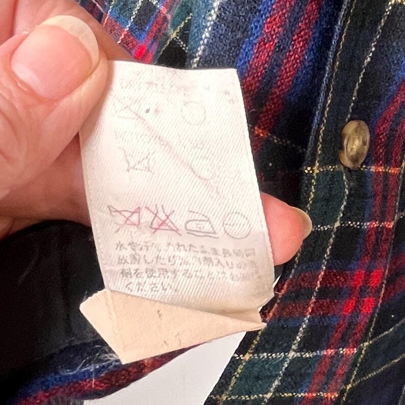 May include: Close-up of a plaid shirt in red, blue, and green. A white tag with care symbols and Japanese text is held up near the shirt. A gold-coloured button is visible on the shirt.