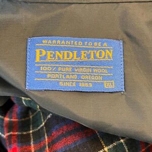 May include: A blue label with gold text reads "PENDLETON" and "100% PURE VIRGIN WOOL." The label also states "PORTLAND, OREGON" and "SINCE 1863." The label is sewn into a dark green fabric, with a plaid pattern visible below.