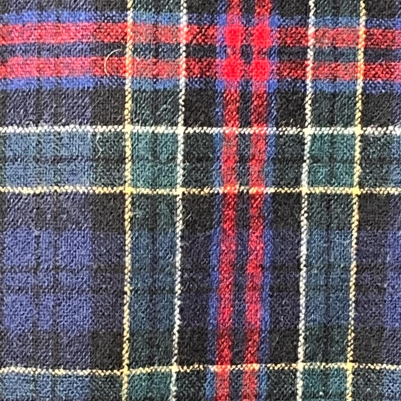 May include: A close-up shot of a tartan fabric. The pattern is a grid of intersecting lines in dark blue, red, green, and yellow. The fabric has a textured surface, possibly wool or a similar woven material.