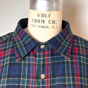 May include: A close-up shot of a plaid shirt with a button-down collar. The shirt has a navy blue, green, red, and yellow plaid pattern. The buttons are visible, and the shirt is displayed on a mannequin.