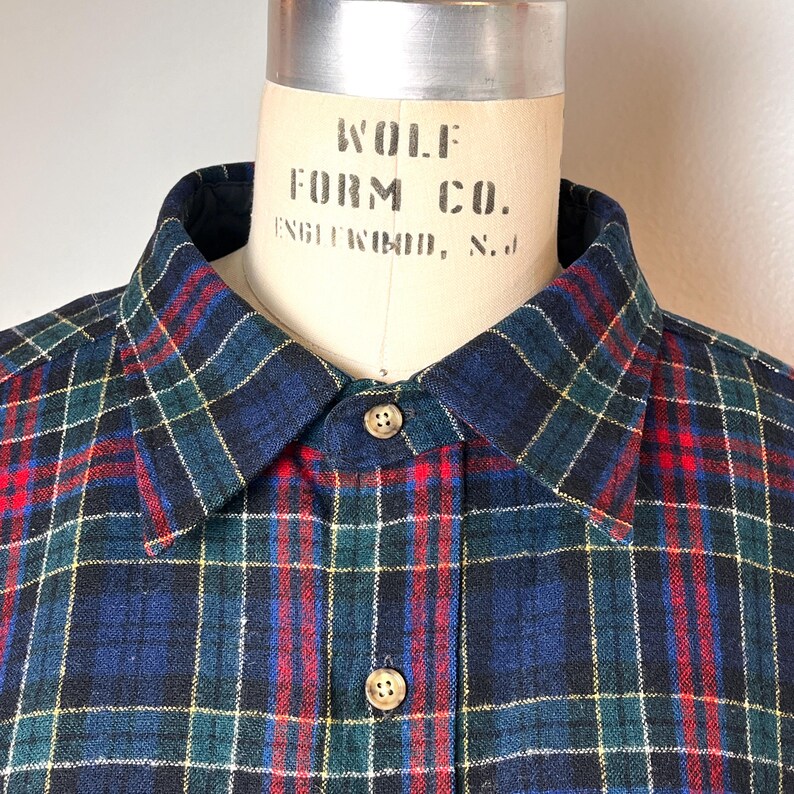 May include: A close-up shot of a plaid shirt with a button-down collar. The shirt has a navy blue, green, red, and yellow plaid pattern. The buttons are visible, and the shirt is displayed on a mannequin.