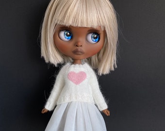 May include: A doll with blonde hair and brown skin wearing a white sweater with a pink heart and a white tulle skirt. The doll is wearing white shoes.