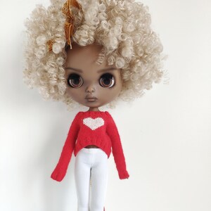 May include: A doll with brown skin, blonde curly hair, and a red sweater with a white heart on the front. The doll is wearing white pants and has a gold bow in her hair.