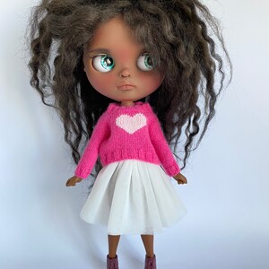 May include: A doll with dark skin, large green eyes, and dark curly hair. The doll wears a pink sweater with a white heart, a white tulle skirt, and burgundy sneakers with white soles.