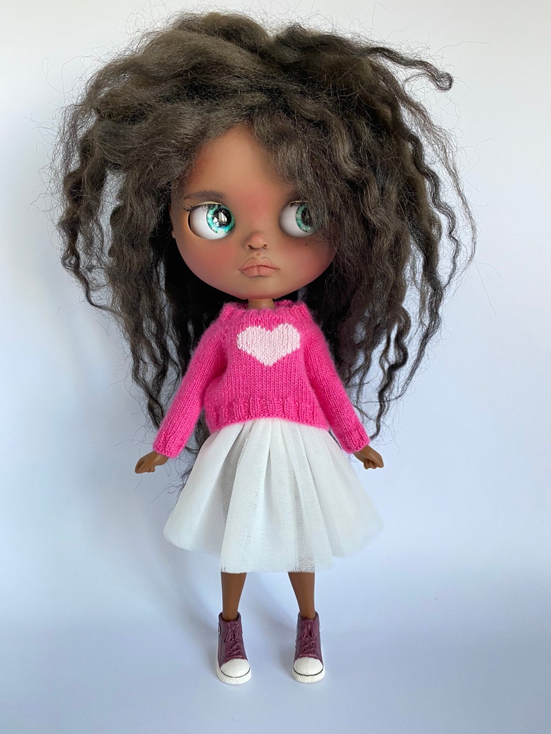 May include: A doll with dark skin, large green eyes, and dark curly hair. The doll wears a pink sweater with a white heart, a white tulle skirt, and burgundy sneakers with white soles.