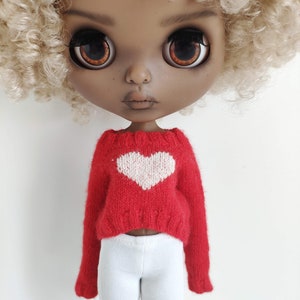 May include: A doll with brown skin and blonde curly hair is wearing a red sweater with a white heart on the front and white pants.