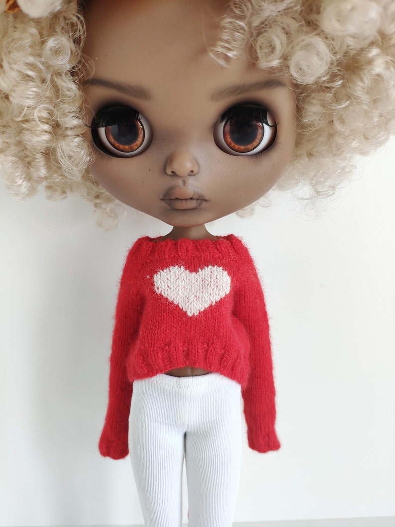 May include: A doll with brown skin and blonde curly hair is wearing a red sweater with a white heart on the front and white pants.