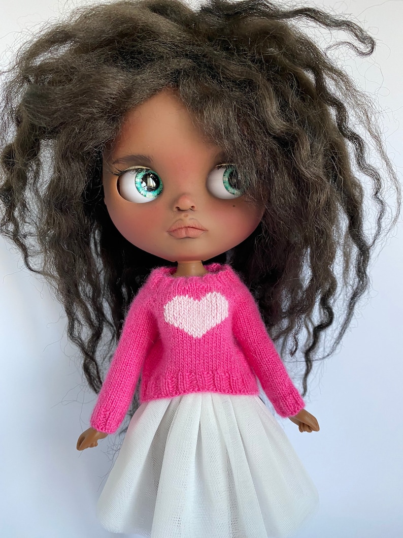 May include: A doll with dark skin, large turquoise eyes, and dark curly hair. The doll wears a pink sweater with a white heart and a white tulle skirt. The doll's arms are visible.