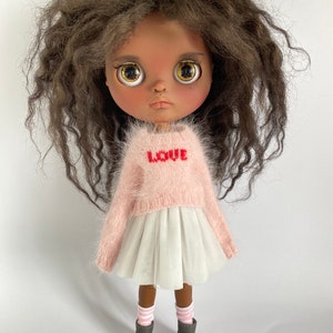 Può includere: Una bambola con capelli castano ricci, che indossa un maglione rosa con la scritta "LOVE", una gonna di tulle bianca, calze a righe rosa e bianche e stivali grigi.