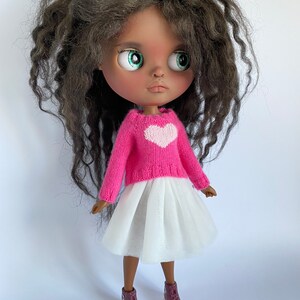 May include: A doll with dark skin, green eyes, and long, wavy brown hair. The doll wears a pink sweater with a white heart, a white tulle skirt, and burgundy sneakers with white soles. The doll is a collectible item.