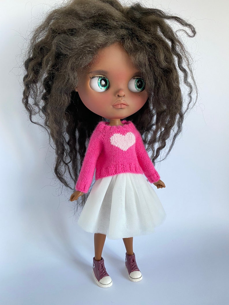 May include: A doll with dark skin, green eyes, and long, wavy brown hair. The doll wears a pink sweater with a white heart, a white tulle skirt, and burgundy sneakers with white soles. The doll is a collectible item.