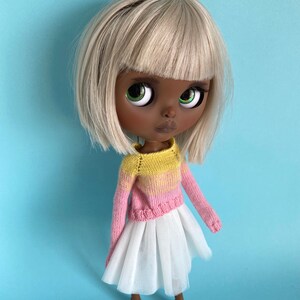 May include: A doll with dark brown skin, blonde hair, and green eyes is wearing a pink and yellow knitted sweater and a white tulle skirt. The doll is standing on a light blue background.