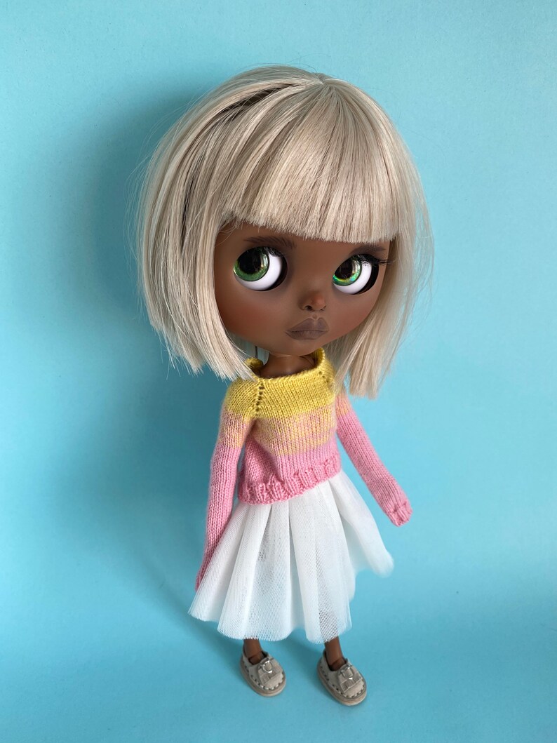 May include: A doll with dark brown skin, blonde hair, and green eyes is wearing a pink and yellow knitted sweater and a white tulle skirt. The doll is standing on a light blue background.