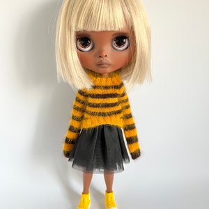 Puede incluir: Una muñeca con cabello rubio que lleva un suéter a rayas amarillas y marrones, una falda de tul negra y zapatillas amarillas.