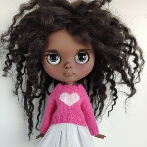 May include: A doll with dark skin, large blue eyes, and dark curly hair. The doll wears a pink sweater with a white heart and a white tulle skirt. The doll is a collectible item.