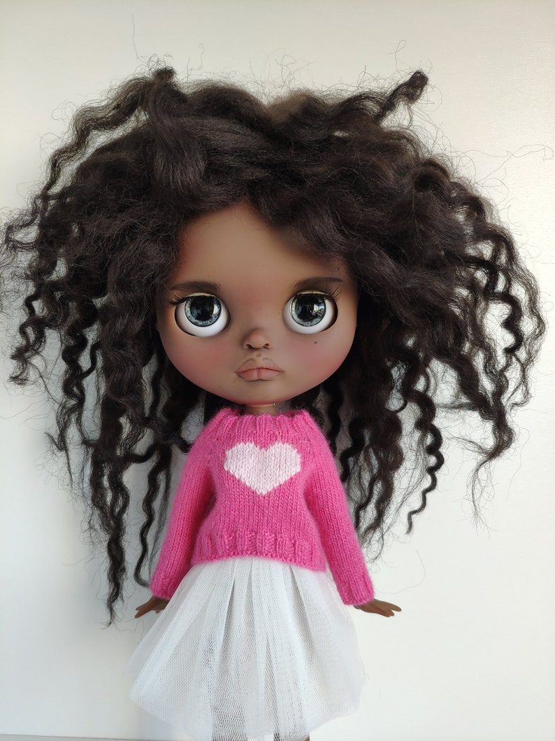May include: A doll with dark skin, large blue eyes, and dark curly hair. The doll wears a pink sweater with a white heart and a white tulle skirt. The doll is a collectible item.