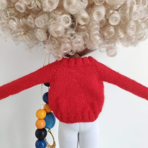 May include: Red knitted sweater with long sleeves for a doll. The sweater is a bright red color and has a ribbed texture.