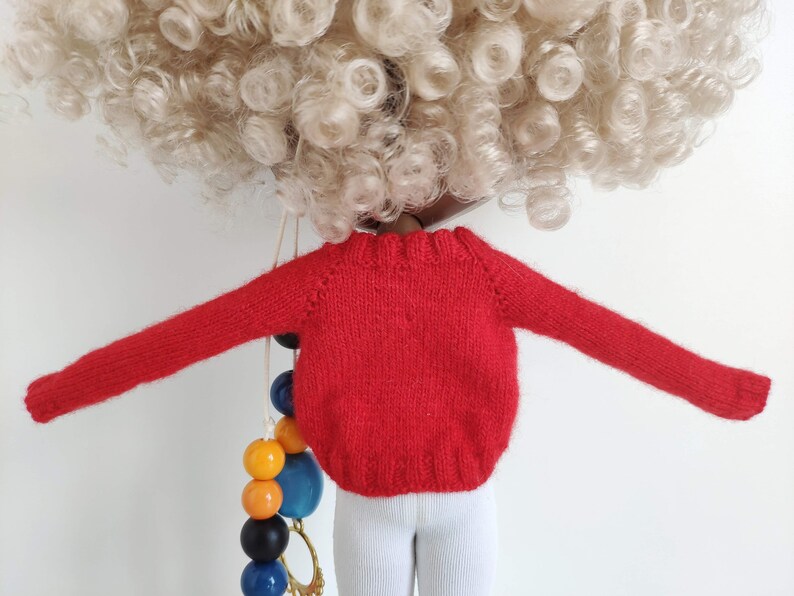 May include: Red knitted sweater with long sleeves for a doll. The sweater is a bright red color and has a ribbed texture.