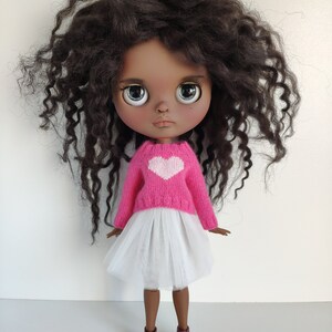 May include: A doll with dark brown curly hair, wearing a pink sweater with a white heart, a white tulle skirt, and burgundy sneakers. The doll has large, expressive eyes and a neutral expression.