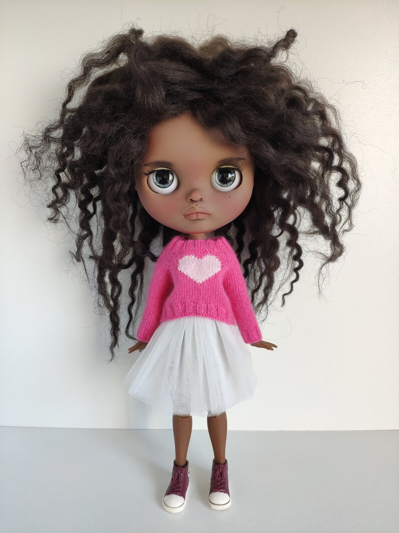 May include: A doll with dark brown curly hair, wearing a pink sweater with a white heart, a white tulle skirt, and burgundy sneakers. The doll has large, expressive eyes and a neutral expression.