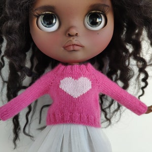 May include: A doll with dark skin, large blue eyes, and curly black hair. The doll wears a pink knitted sweater with a white heart design and a white tulle skirt. The doll's arms are outstretched.