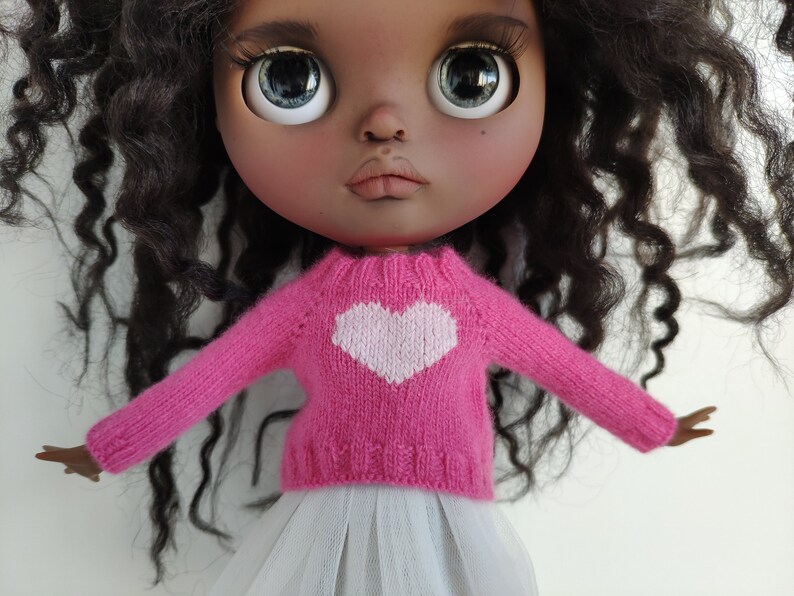May include: A doll with dark skin, large blue eyes, and curly black hair. The doll wears a pink knitted sweater with a white heart design and a white tulle skirt. The doll's arms are outstretched.