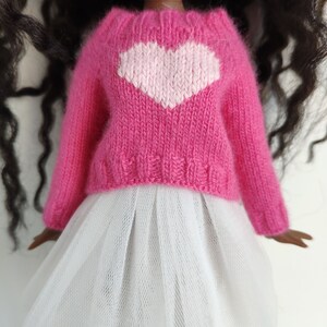 May include: A doll wearing a pink knitted sweater with a white heart design and a white tulle skirt. The sweater has long sleeves and a textured knit pattern. The doll has dark hair and a fair complexion.