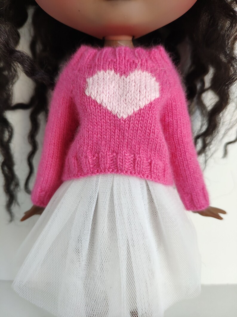 May include: A doll wearing a pink knitted sweater with a white heart design and a white tulle skirt. The sweater has long sleeves and a textured knit pattern. The doll has dark hair and a fair complexion.
