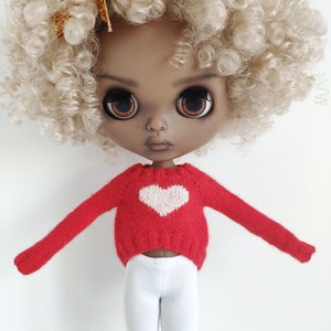 May include: A doll with a red sweater with a white heart on the front, white pants, and blonde curly hair.