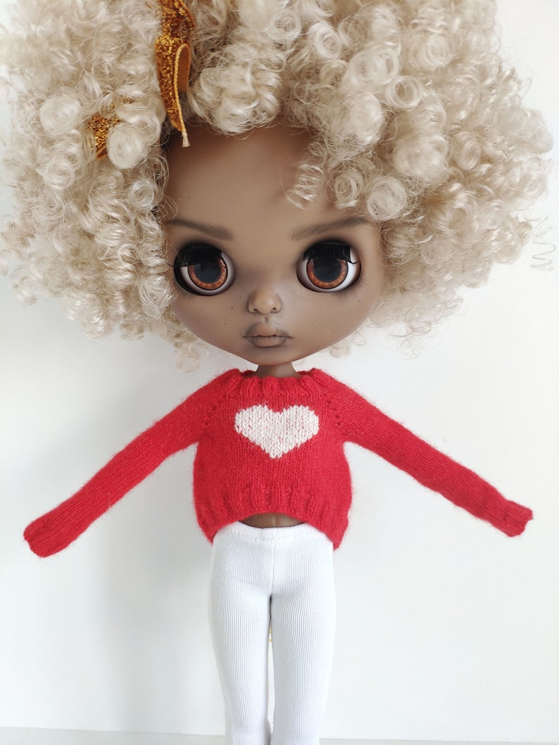 May include: A doll with a red sweater with a white heart on the front, white pants, and blonde curly hair.