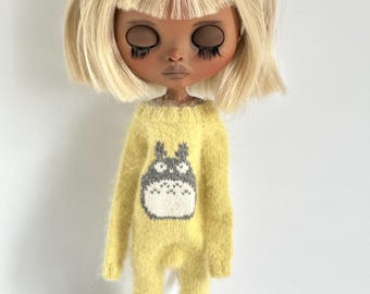 Op de afbeelding: Een pop met blond haar en bruine huid die een gele gebreide jumpsuit draagt met een grijs en wit cartoonfiguur op de voorkant. De pop draagt blauwe schoenen.