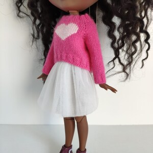 May include: A doll wearing a pink sweater with a white heart design, a white tulle skirt, and burgundy high-top sneakers with white soles. The doll has dark skin and curly black hair.
