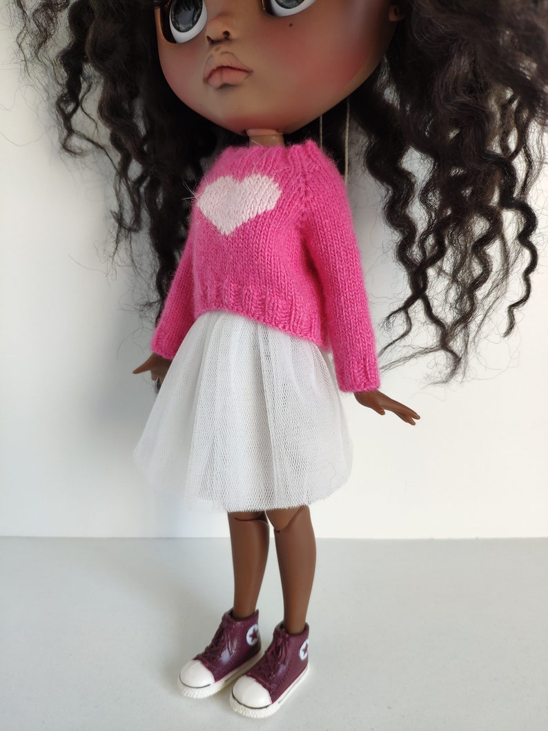 May include: A doll wearing a pink sweater with a white heart design, a white tulle skirt, and burgundy high-top sneakers with white soles. The doll has dark skin and curly black hair.
